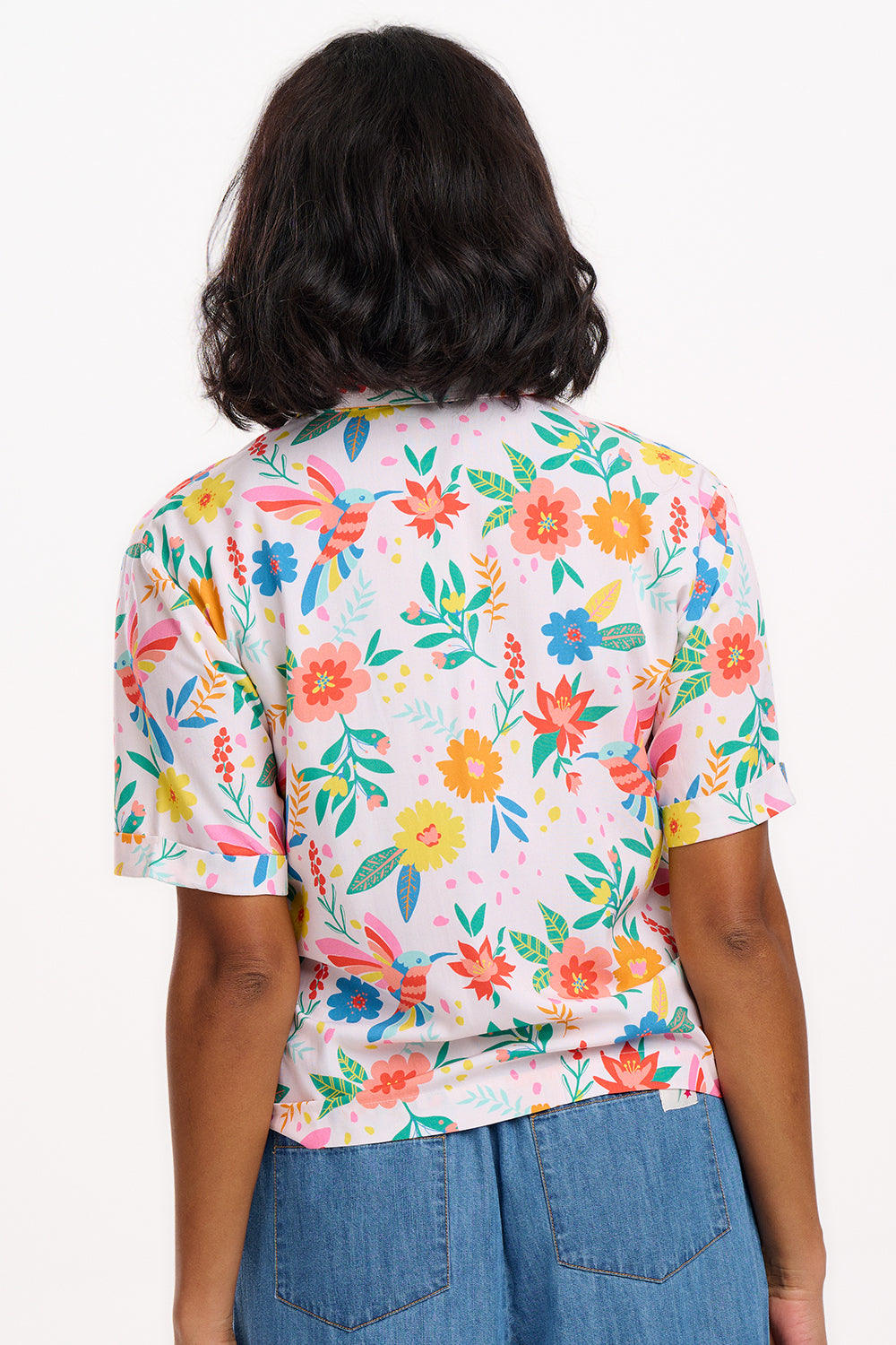 A woman wearing an off-white shirt with a tropical hummingbird print, short sleeves, and rolled-up cuffs.