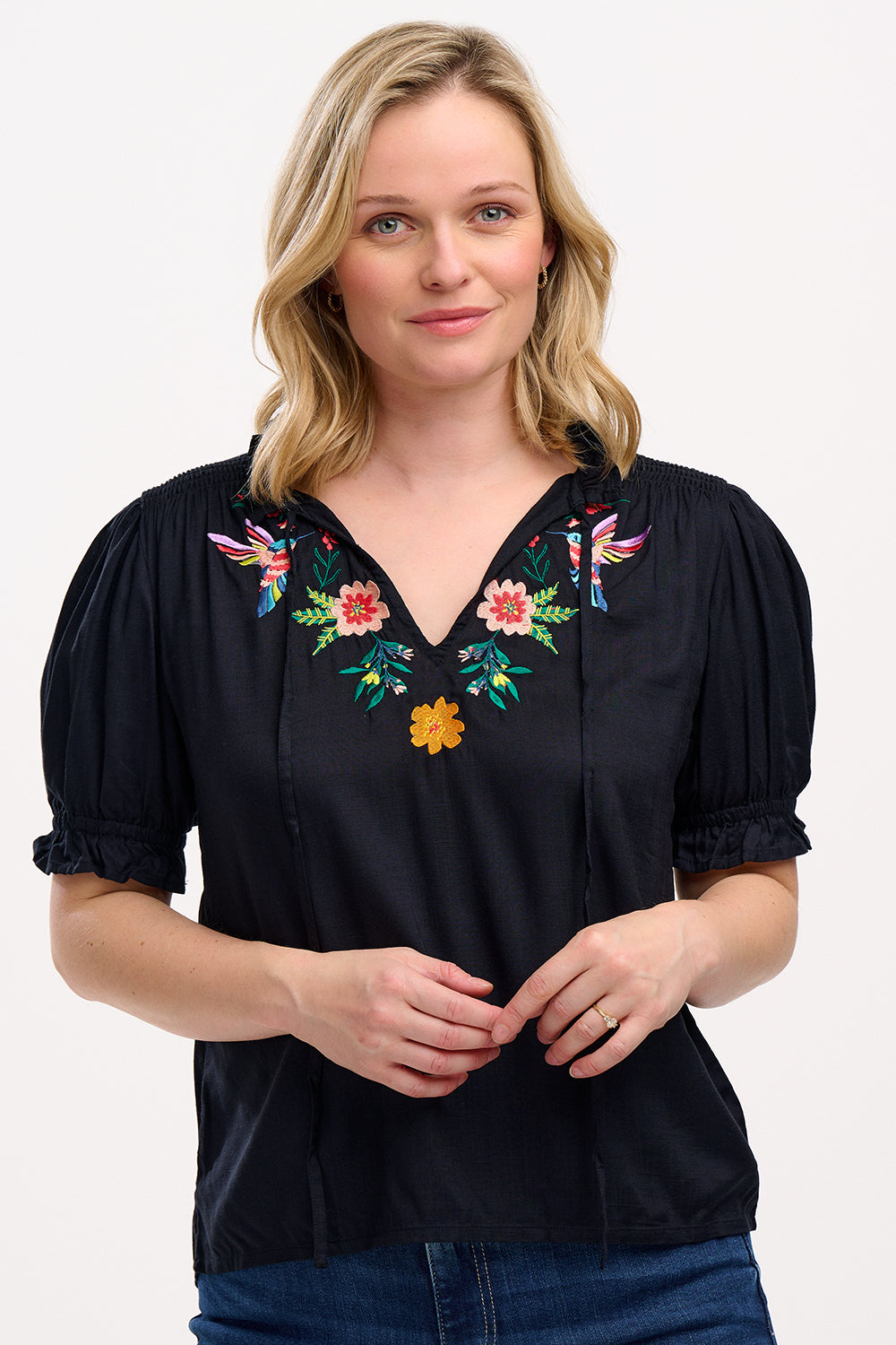 A woman wearing a black Angelique blouse with floral embroidery, short frill sleeves, and a V-neckline.