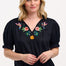 A woman wearing a black Angelique blouse with floral embroidery, short frill sleeves, and a V-neckline.