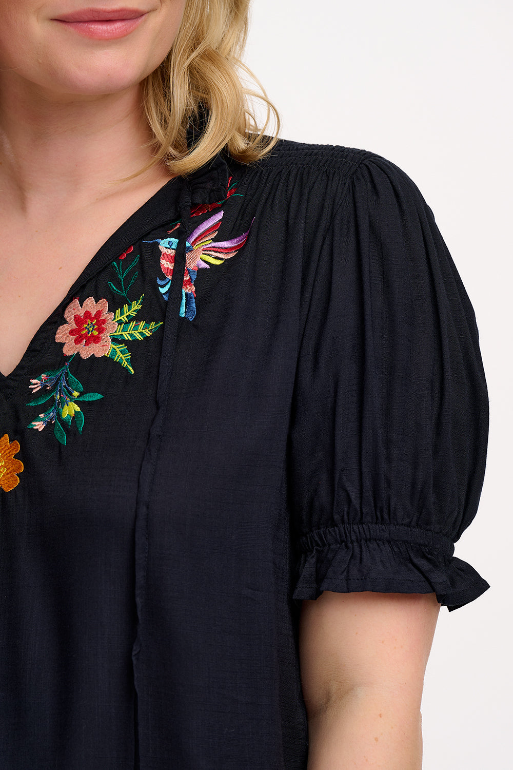 A woman wearing a black Angelique blouse with floral embroidery, short frill sleeves, and a V-neckline.