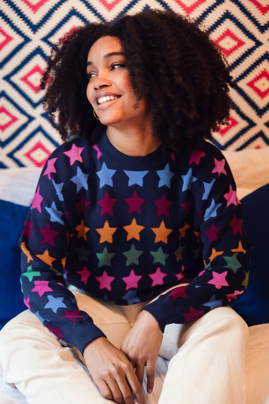 A woman smiling while wearing a multicolored star-patterned jumper with a round neckline and long sleeves.
