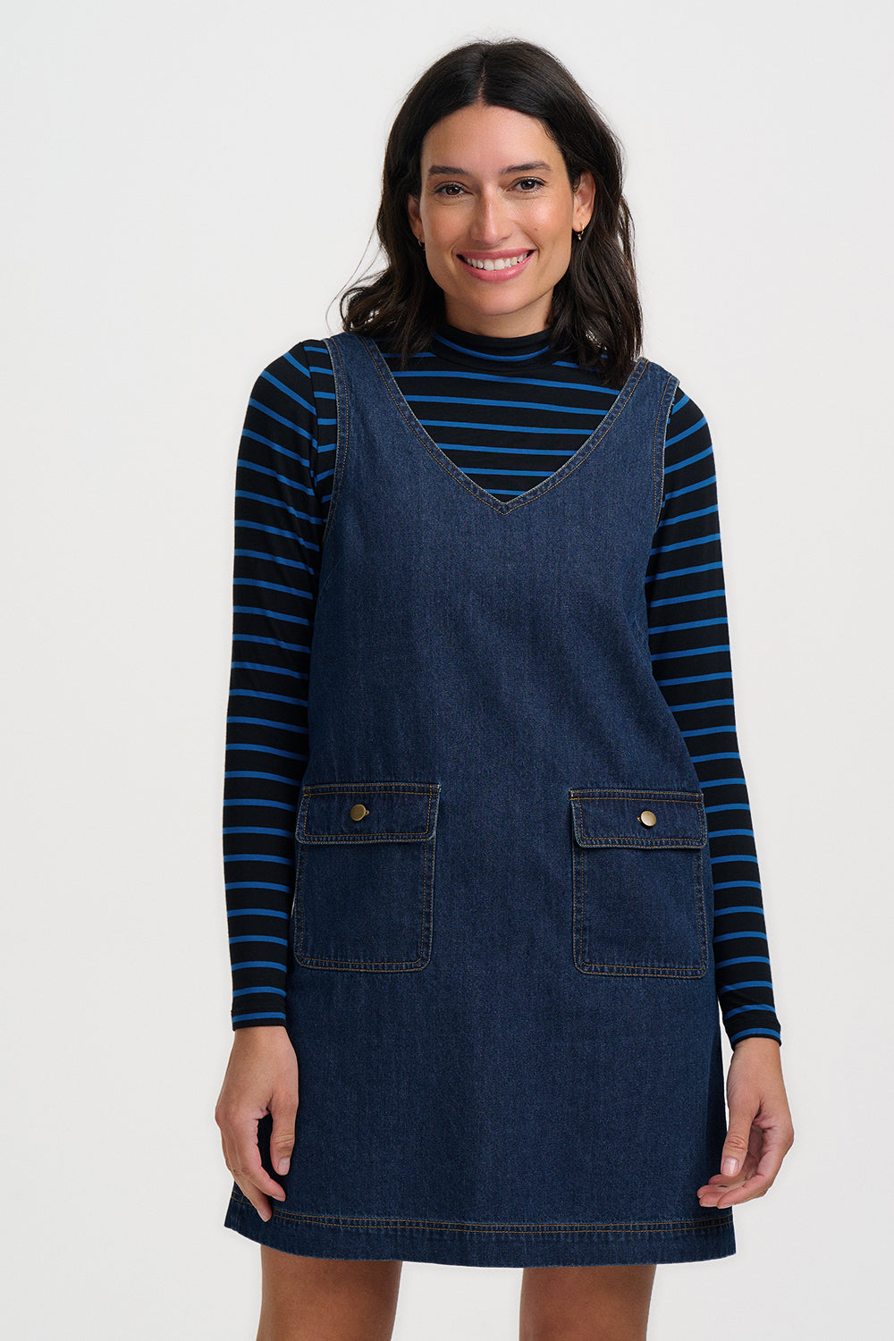 A woman modeling a dark blue denim pinafore dress layered over a navy and navy blue striped long-sleeve turtleneck, set against a plain light gray studio background.