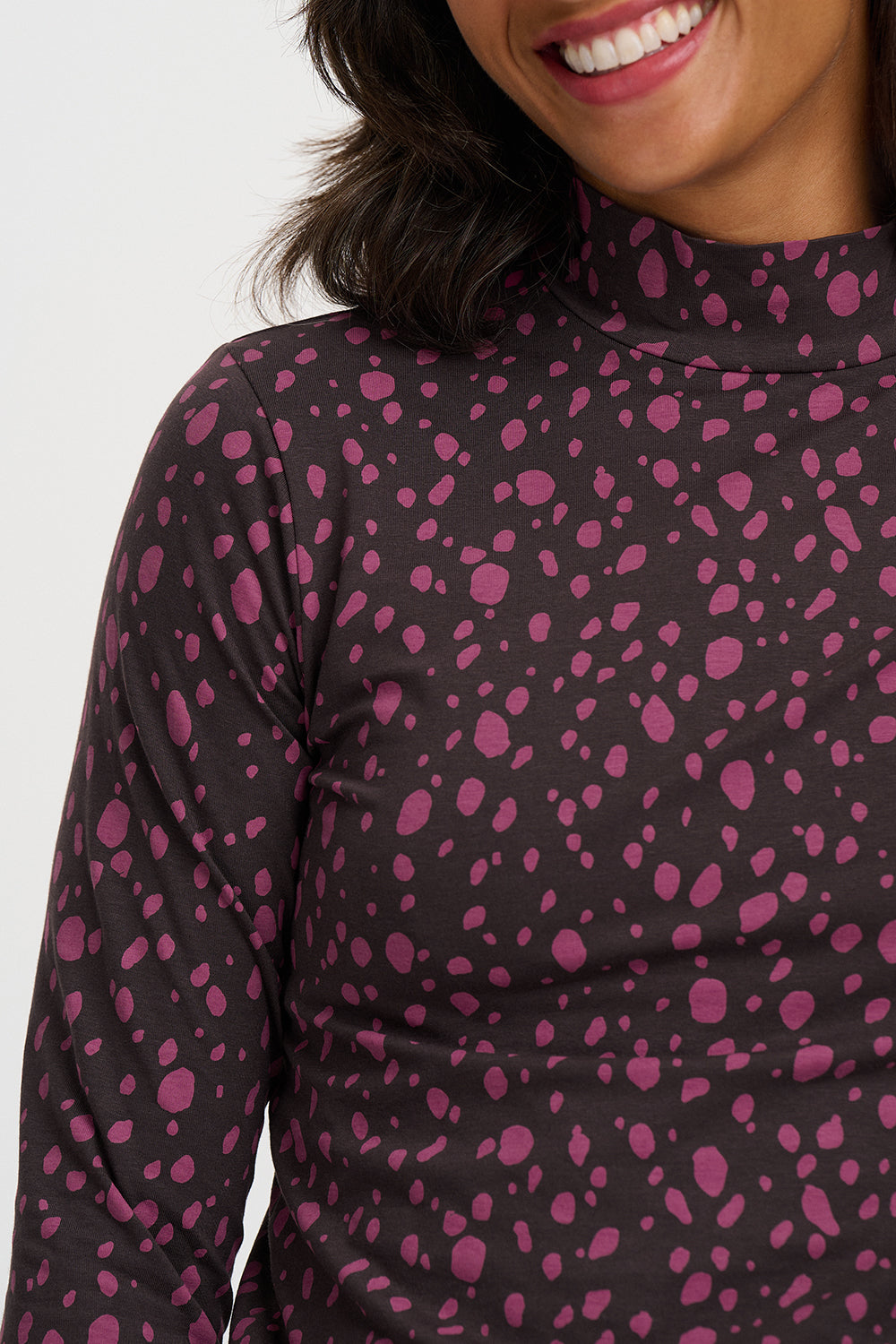 Close-up of a woman wearing a long-sleeve mock neck top with a dark charcoal base and magenta pink abstract speckled pattern.