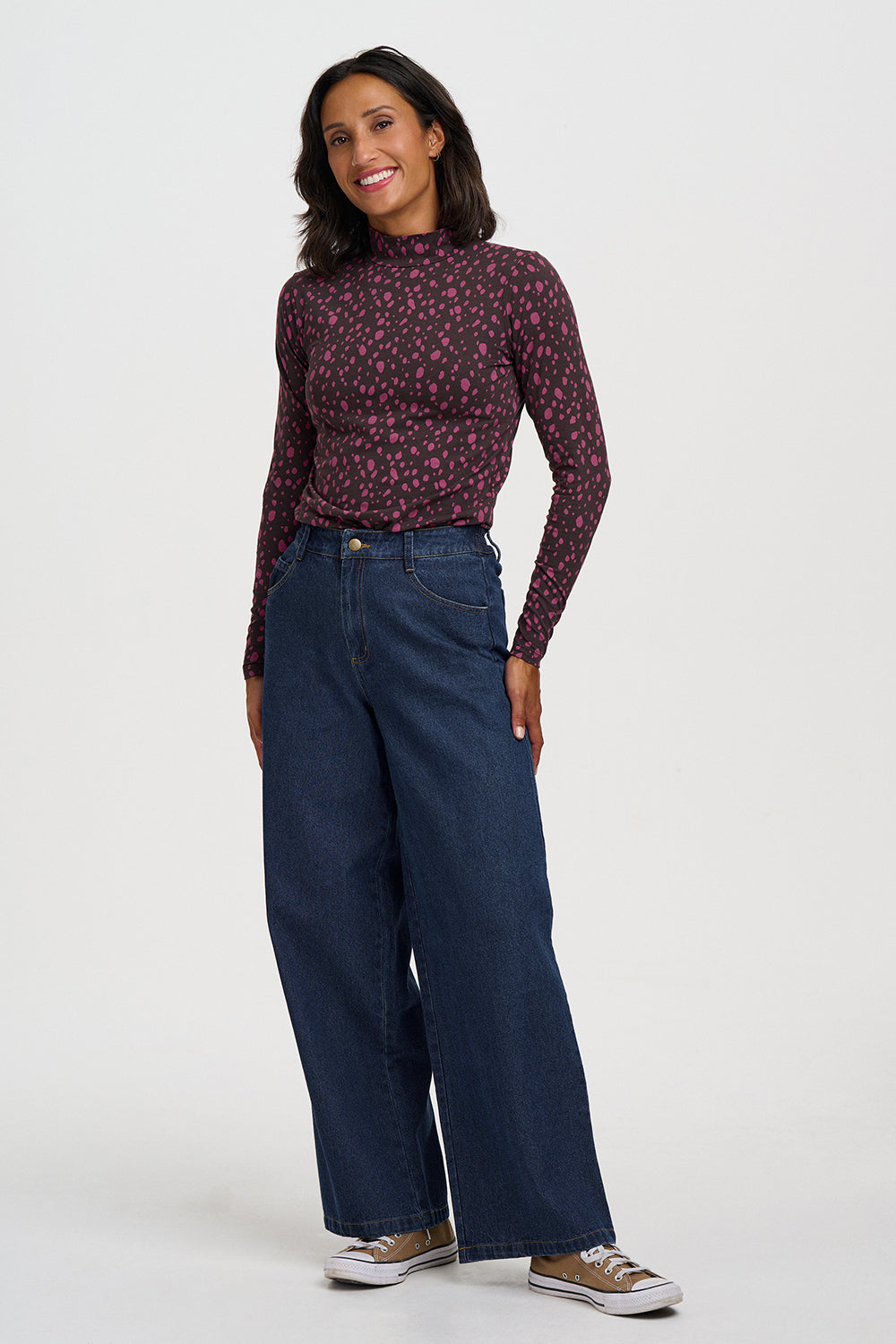 A woman modeling a dark burgundy long-sleeve mock-neck top with a small pink floral pattern, paired with dark blue wide-leg jeans and brown sneakers, against a plain light gray studio background.