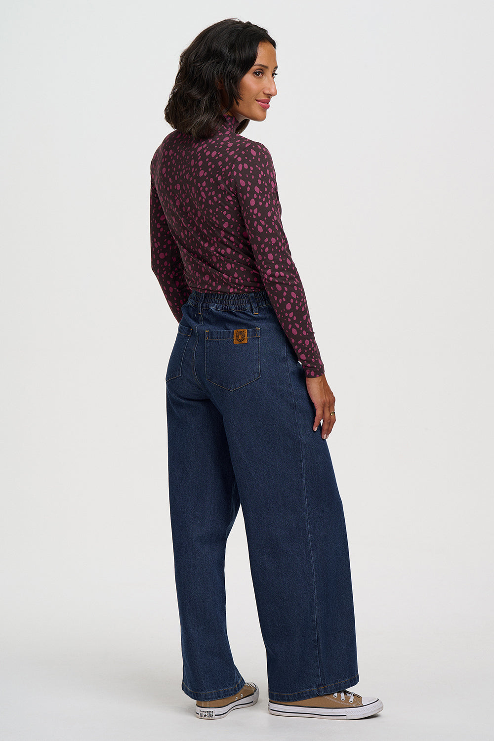 A woman is shown from the back wearing a dark red patterned long-sleeve top and dark blue wide-leg jeans, paired with brown and white sneakers, against a light gray studio background.
