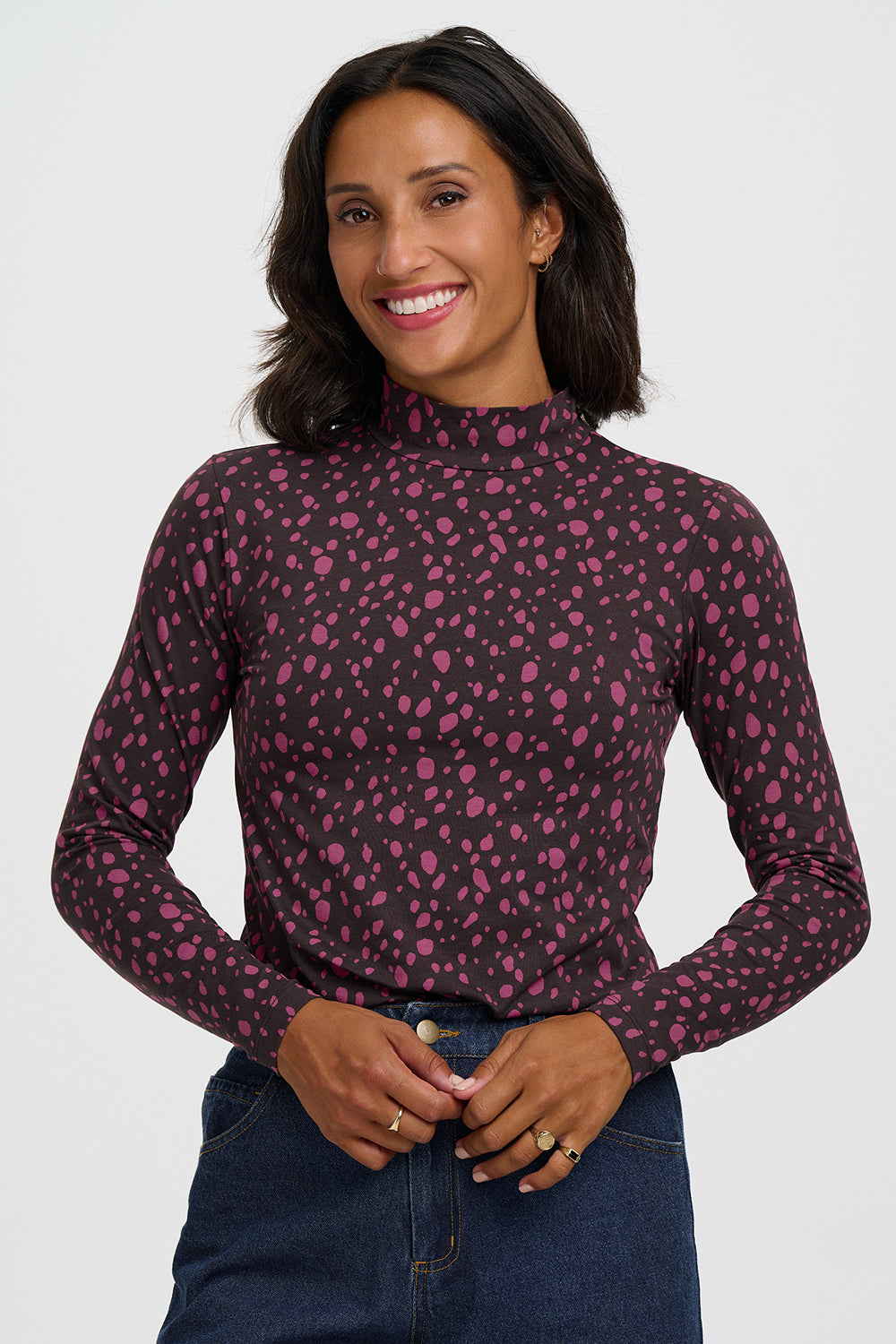 A woman wearing a dark brown mock neck long-sleeve top with a pink leopard print pattern, paired with blue jeans, against a light gray studio background.