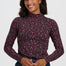 A woman wearing a dark brown mock neck long-sleeve top with a pink leopard print pattern, paired with blue jeans, against a light gray studio background.
