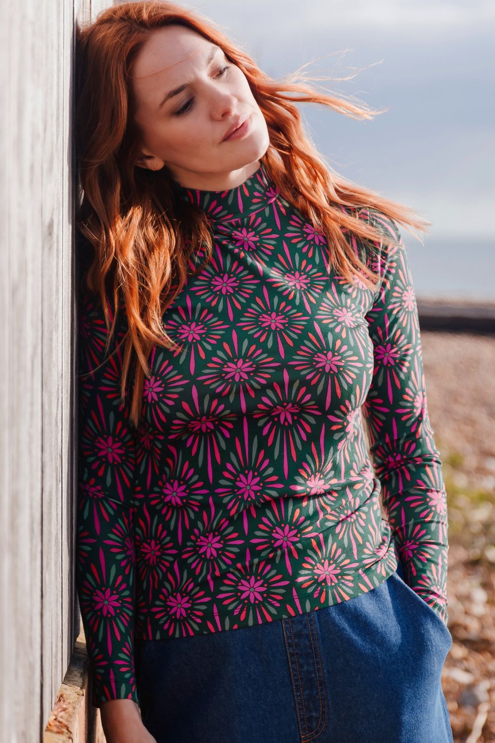 Woman wearing a patterned long-sleeve shirt leaning against a wooden post outdoors.