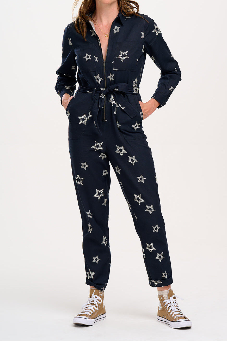 A model wearing a black boilersuit with white triple star patterns on the sleeves and legs, paired with gold sneakers.