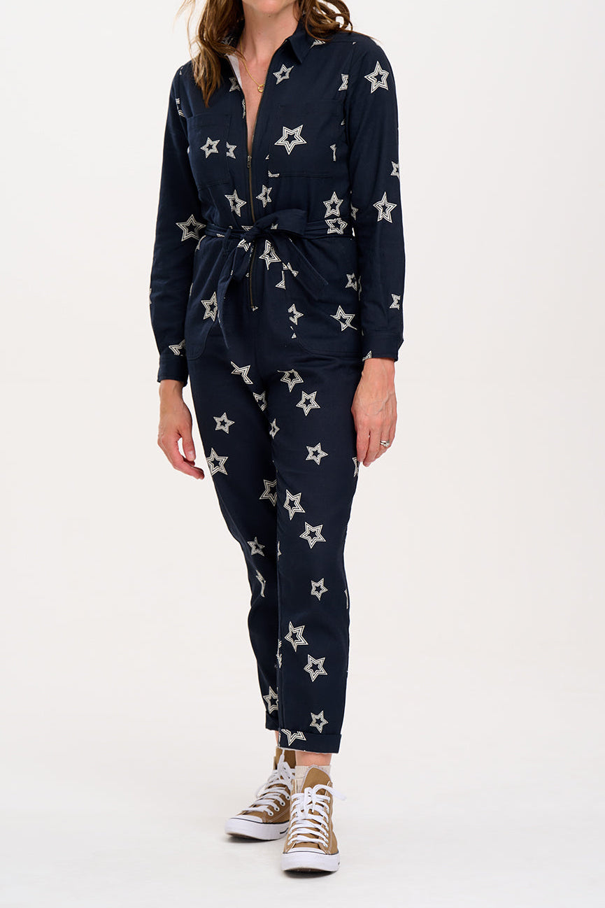 A woman standing in a boilersuit with a black base and white star pattern, featuring a full sleeve shirt upper, exposed front zip, and side pockets. 