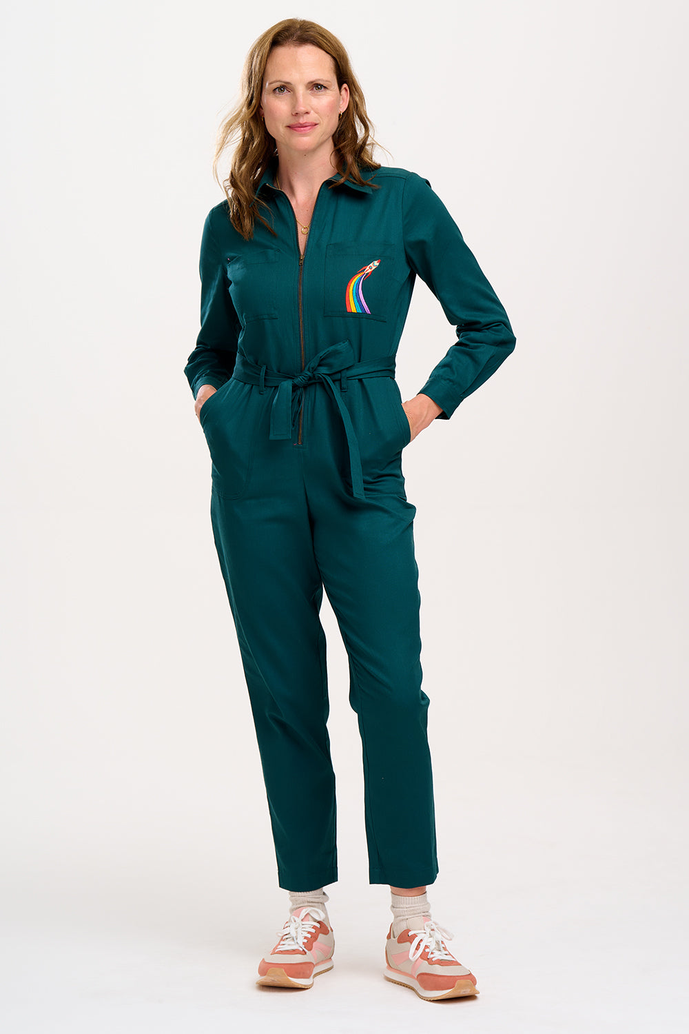 A woman wearing a green boilersuit with a rocket pocket embroidery, paired with white sneakers.