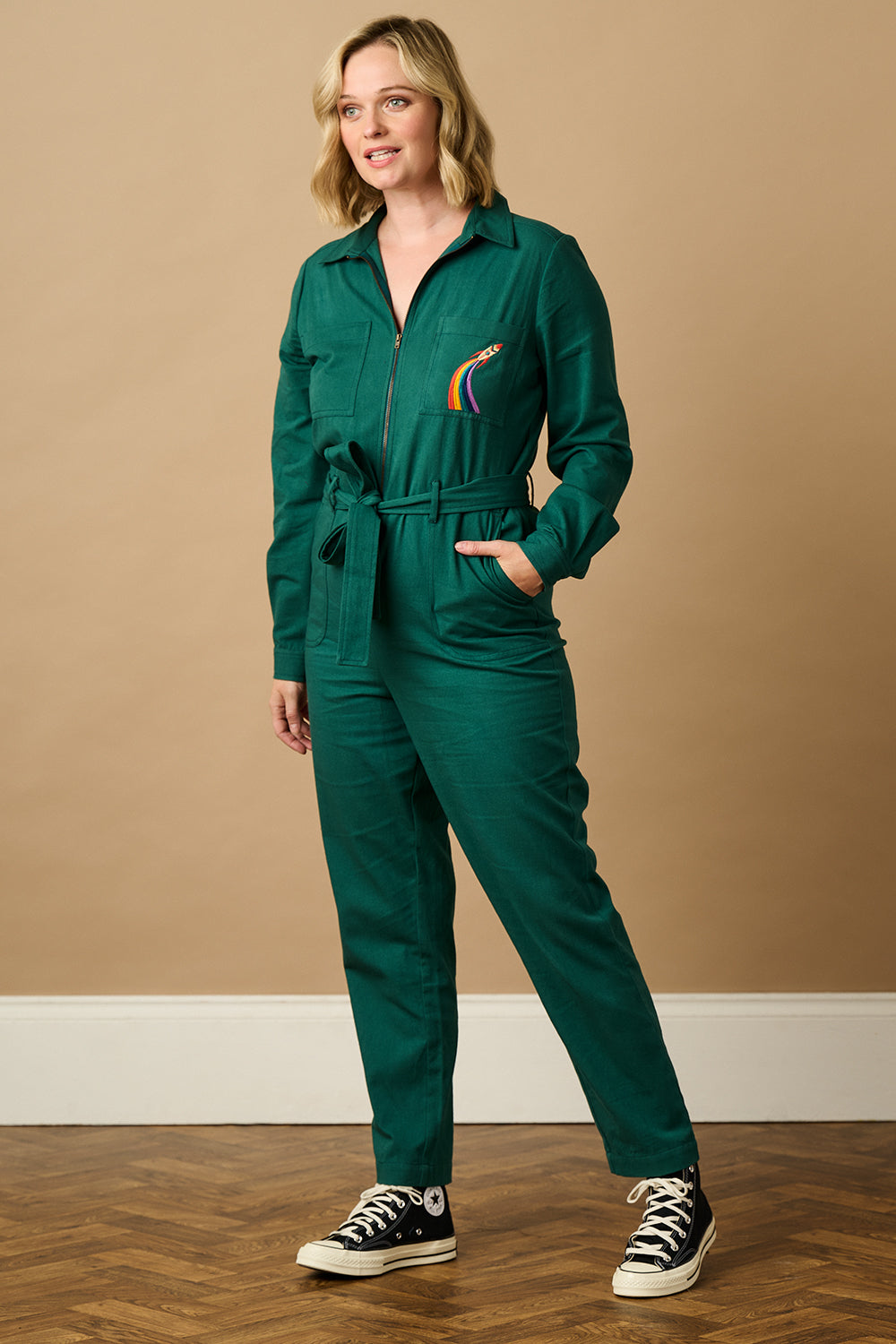 A woman wearing a green boilersuit with a rocket pocket embroidery, paired with white sneakers.