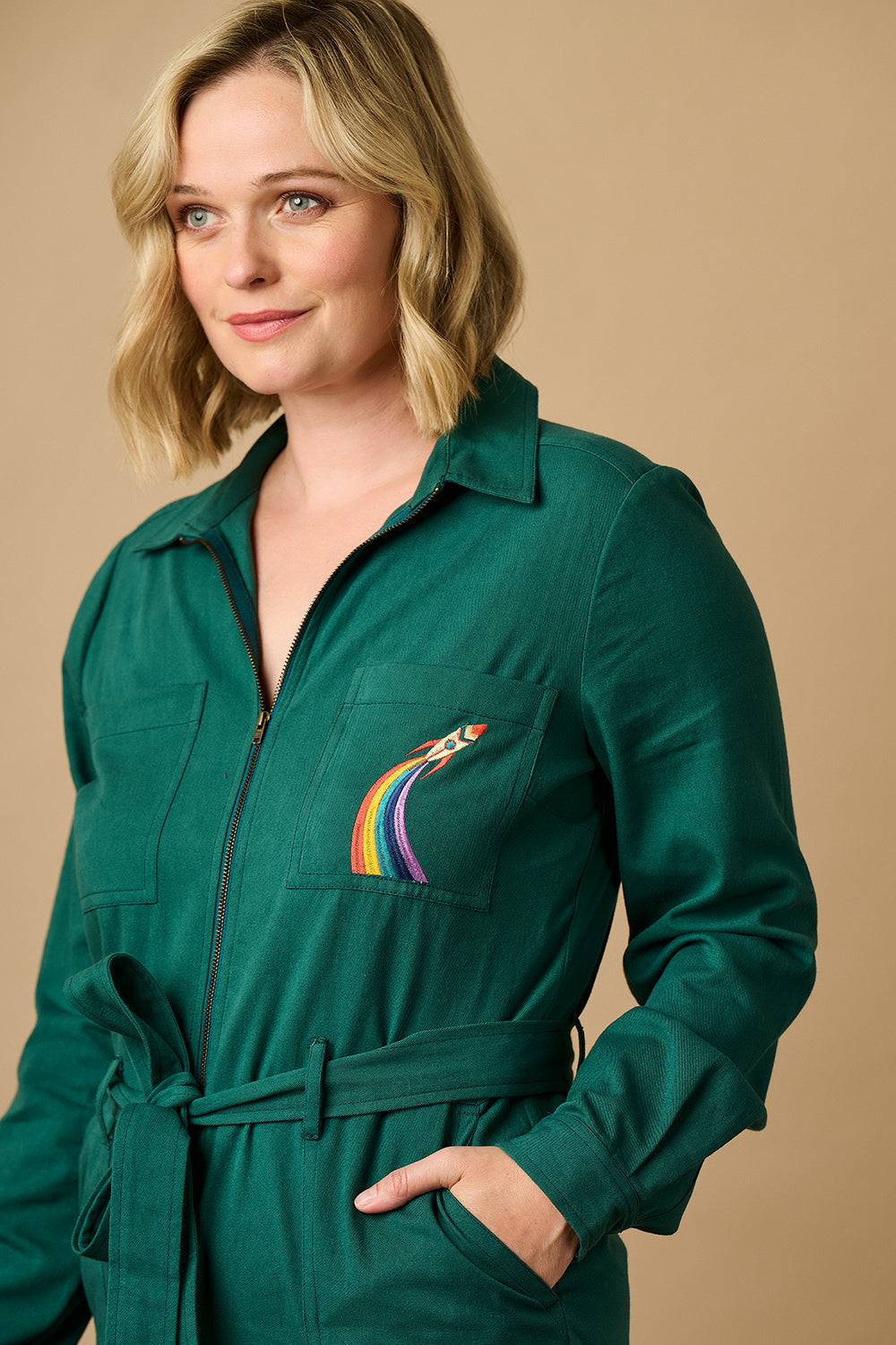 A woman wearing a green boilersuit with a rocket pocket embroidery, paired with white sneakers.