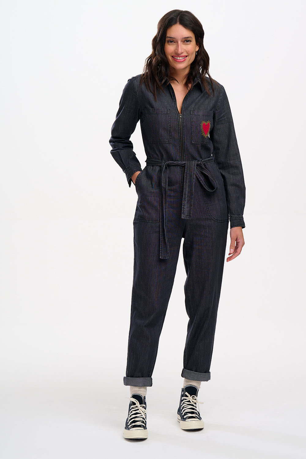 A female model wearing a washed black denim boilersuit with an exposed front zip and embroidered heart on the chest pocket, paired with sneakers.