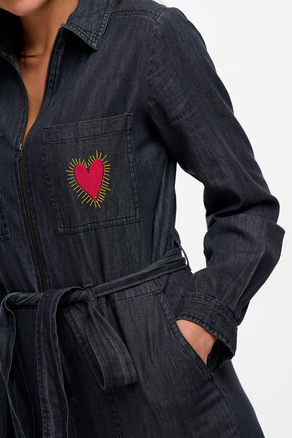 A female model wearing a washed black denim boilersuit with an exposed front zip and embroidered heart on the chest pocket, paired with sneakers.