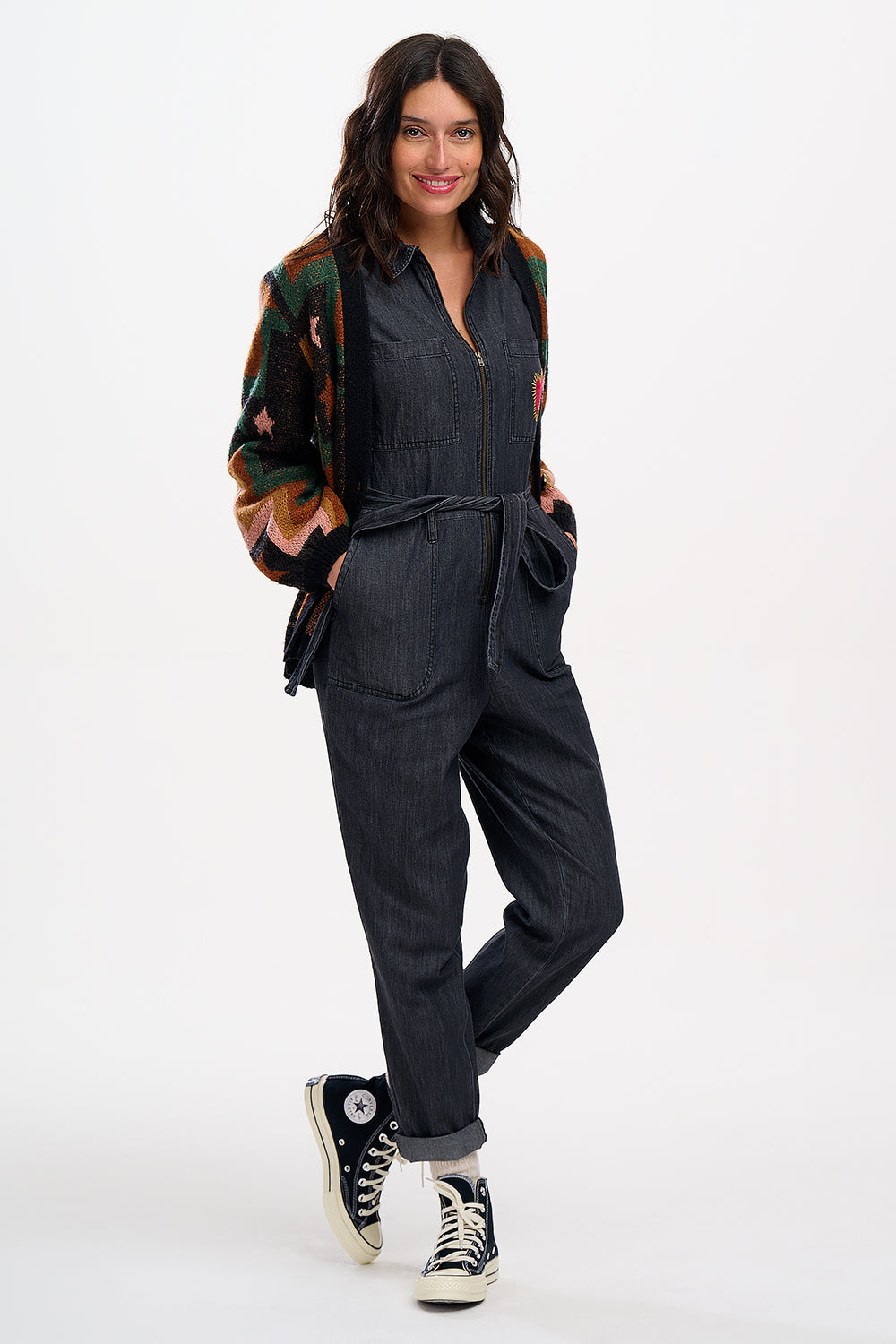 A female model wearing a washed black denim boilersuit with an exposed front zip and embroidered heart on the chest pocket, paired with sneakers.