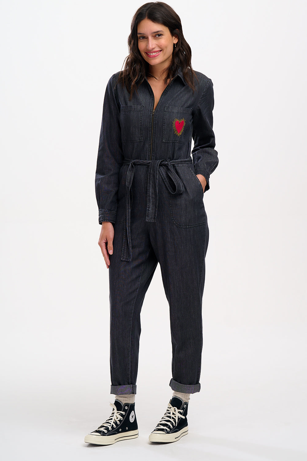 A female model wearing a washed black denim boilersuit with an exposed front zip and embroidered heart on the chest pocket, paired with sneakers.