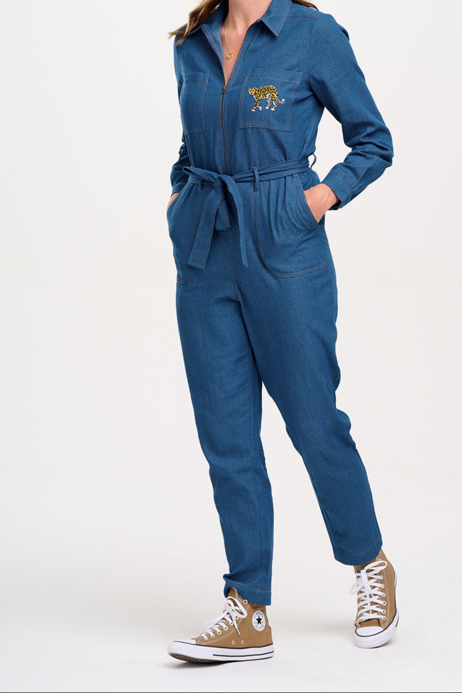 A mid blue denim jumpsuit with a full sleeve shirt upper, an exposed front zip, side pockets, and straight legs. The jumpsuit features an embroidered leopard on the chest pocket.