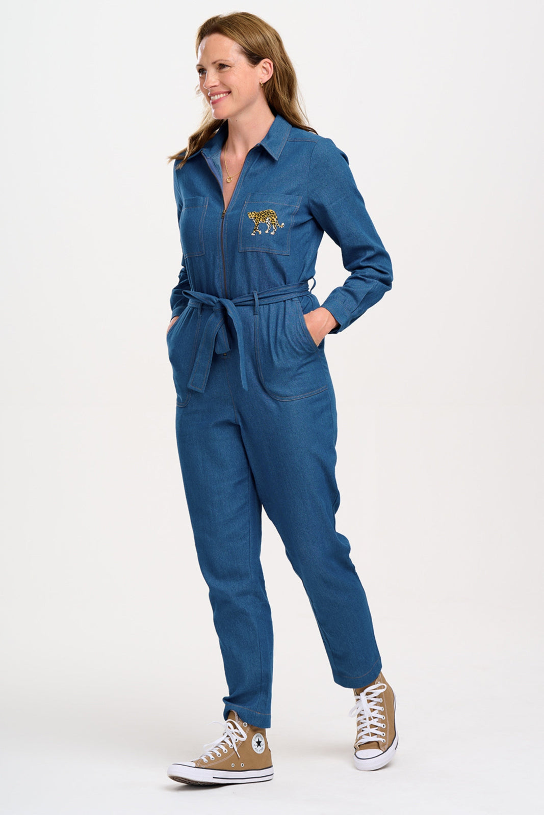 A mid blue denim jumpsuit with a full sleeve shirt upper, an exposed front zip, side pockets, and straight legs. The jumpsuit features an embroidered leopard on the chest pocket.