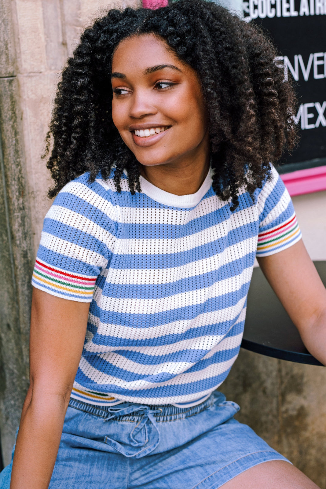 A woman wearing a blue and white striped knit top with rainbow tipping on the cuffs and hem, paired with a denim skirt.