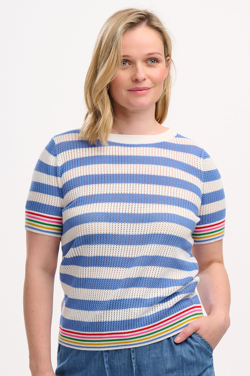 A woman wearing a blue and white striped knit top with rainbow tipping on the cuffs and hem, paired with a denim skirt.