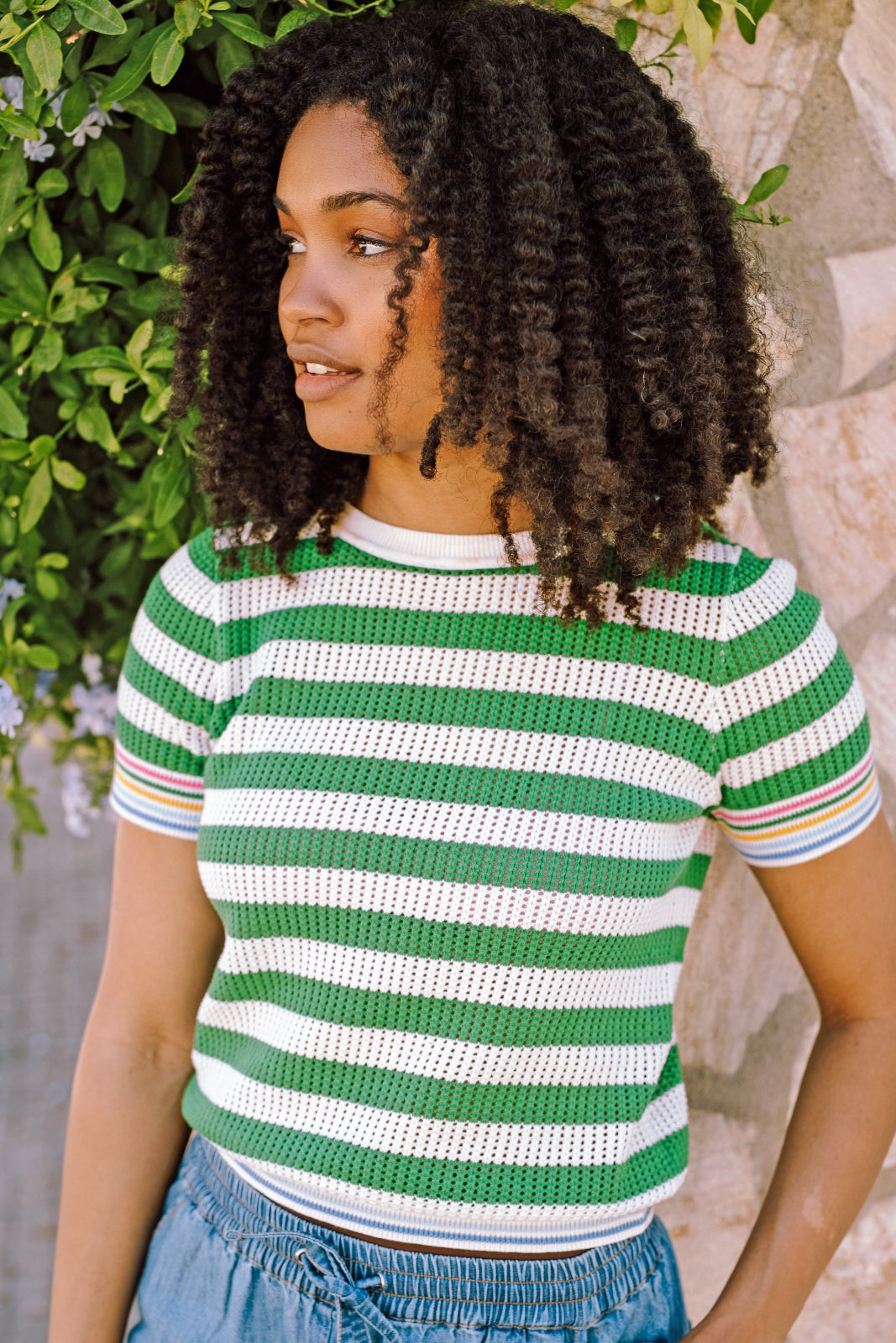 A woman wearing a green and white striped knit top with a crew neck and rainbow tipping on the cuffs and hem.