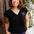 Woman wearing a black t-shirt with a logo and colorful pants, standing against a wooden wall with plants in the background.