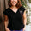 Woman wearing a black t-shirt with a logo and colorful pants, standing against a wooden wall with plants in the background.