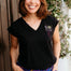 Woman wearing a black t-shirt with a logo and colorful pants, standing against a wooden wall with plants in the background.