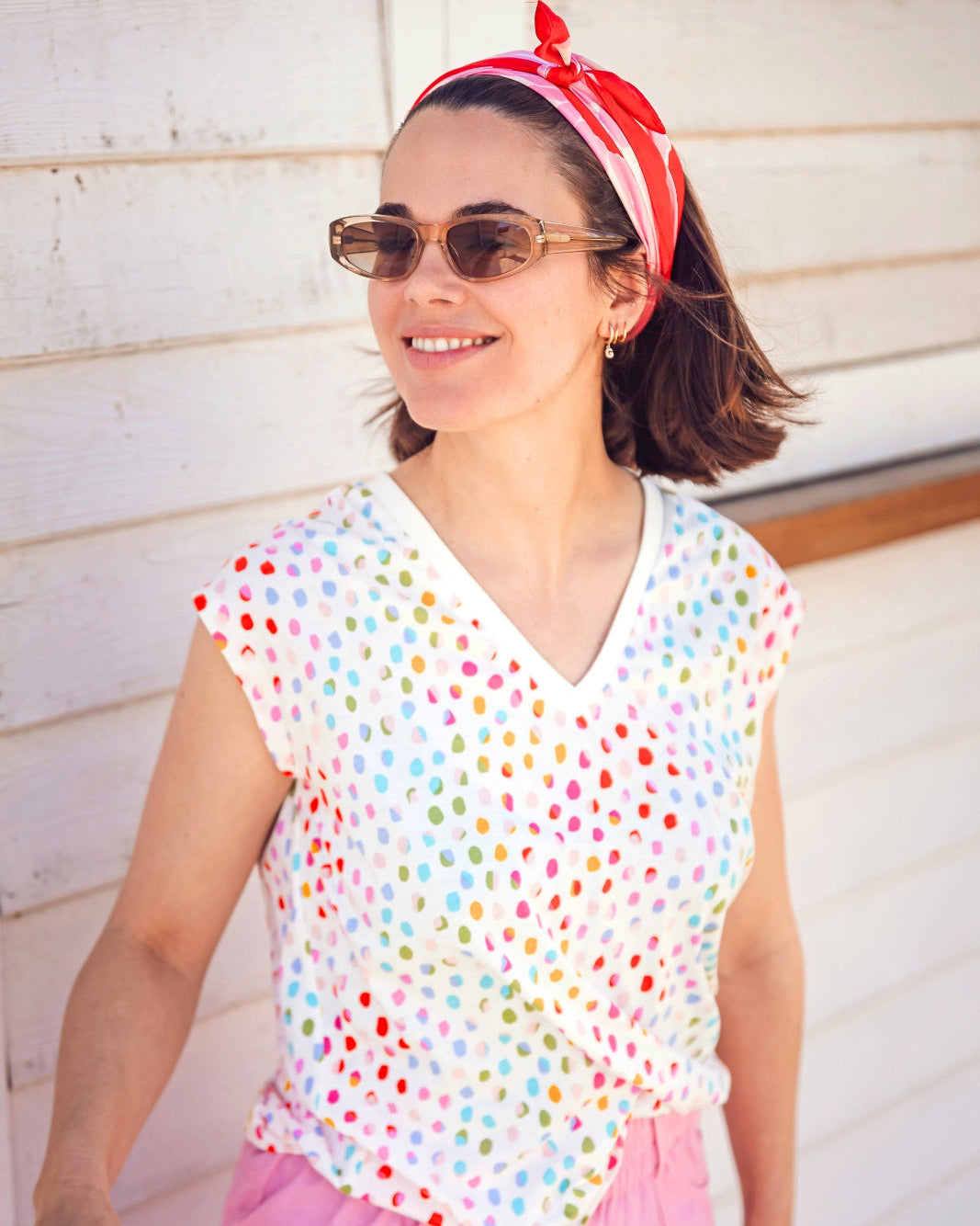 A woman wearing a white V-neck tank t-shirt with a rainbow pebble print, paired with pink pants and accessorized with a red bandana and sunglasses.