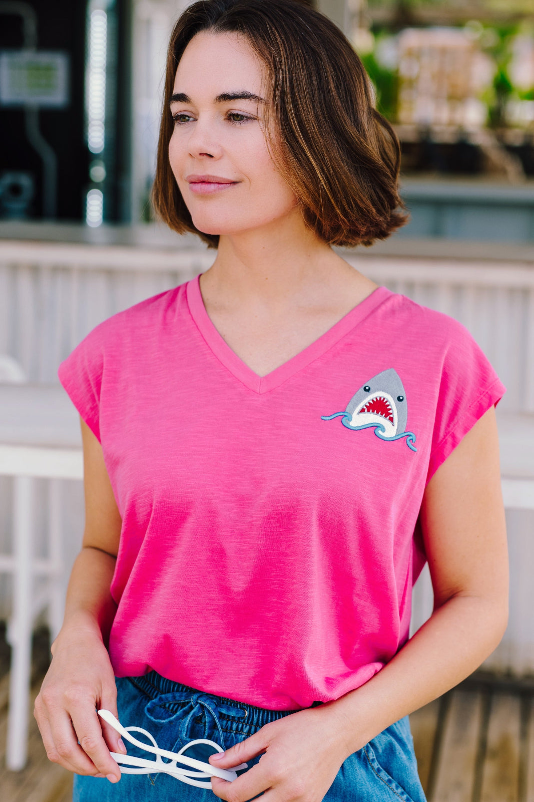 A woman wearing a pink V-neck tank t-shirt with a playful embroidered shark detail, paired with blue denim shorts.