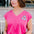 A woman wearing a pink V-neck tank t-shirt with a playful embroidered shark detail, paired with blue denim shorts.