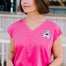 A woman wearing a pink V-neck tank t-shirt with a playful embroidered shark detail, paired with blue denim shorts.