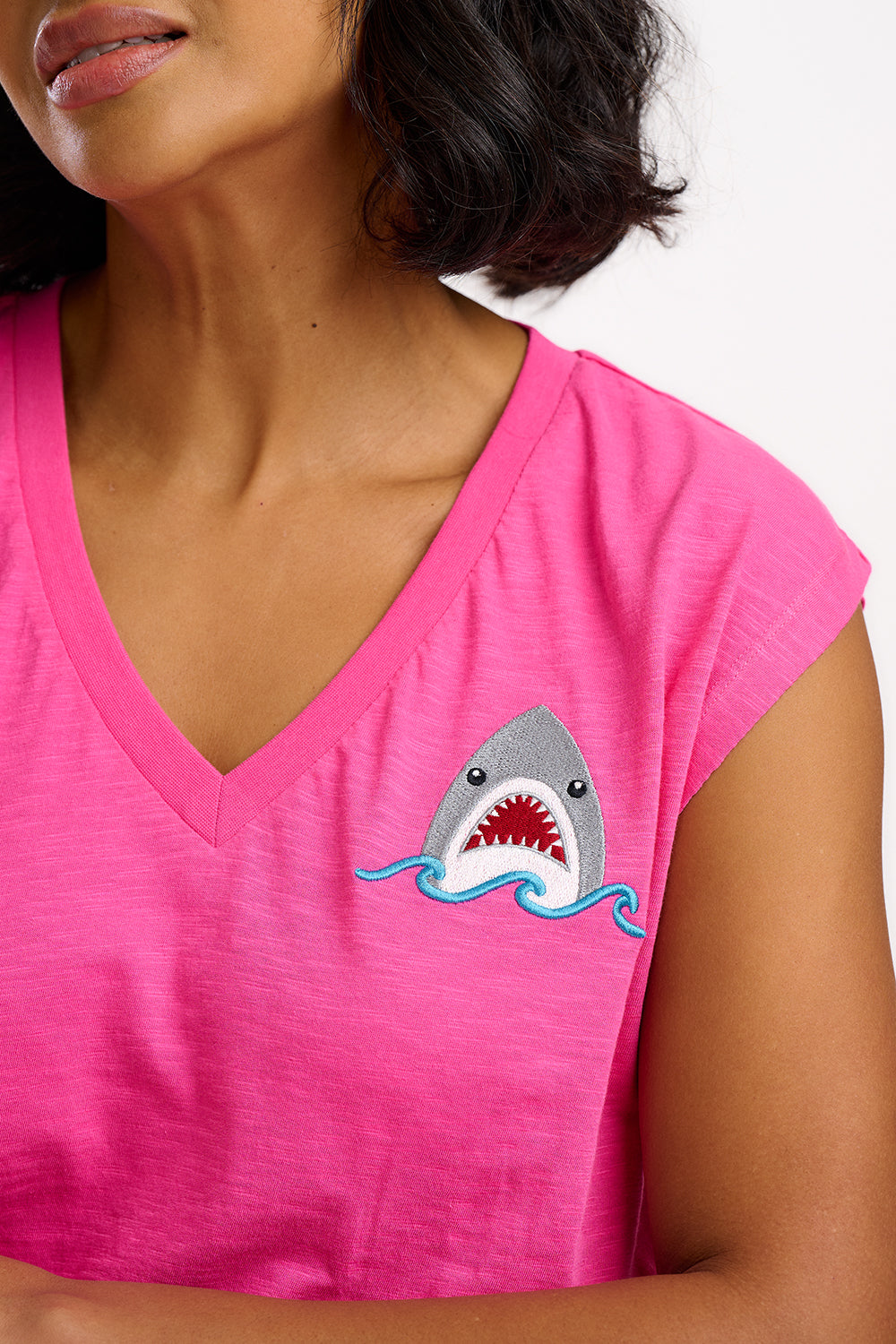 A woman wearing a pink V-neck tank t-shirt with a playful embroidered shark detail