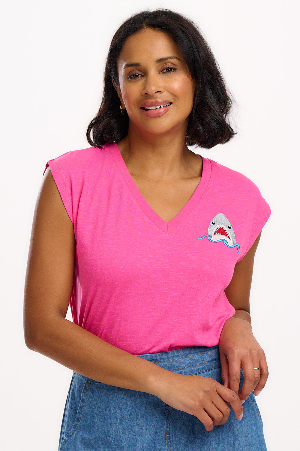 A woman wearing a pink V-neck tank t-shirt with a shark embroidery on the front, paired with a blue skirt.