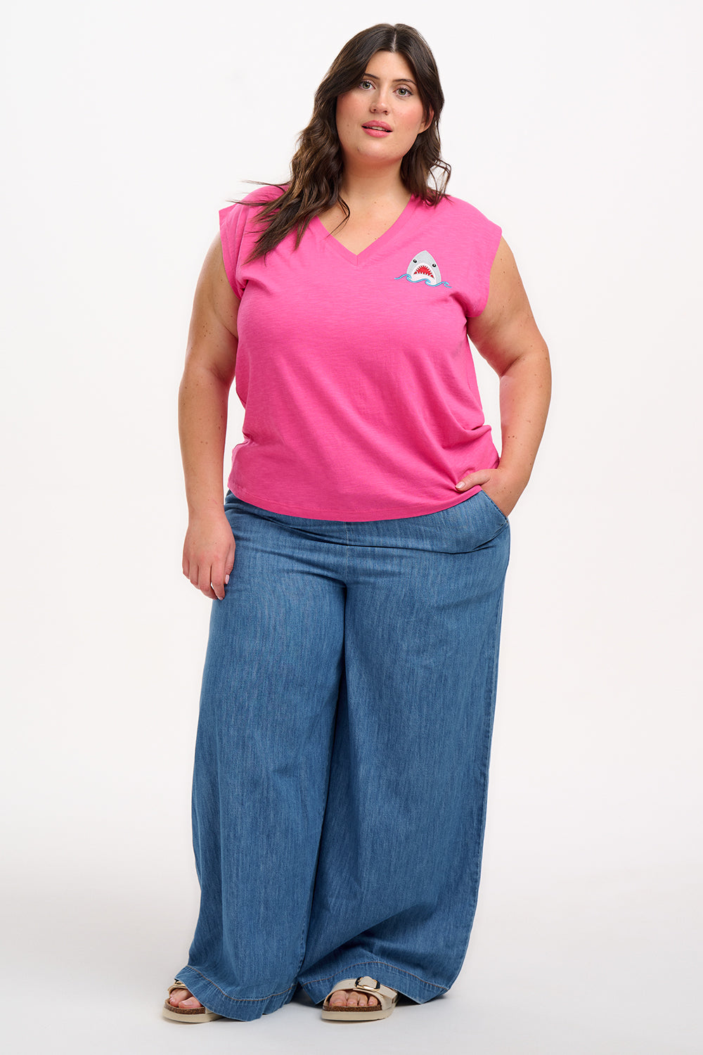 A woman wearing a pink V-neck tank t-shirt with a playful embroidered shark detail