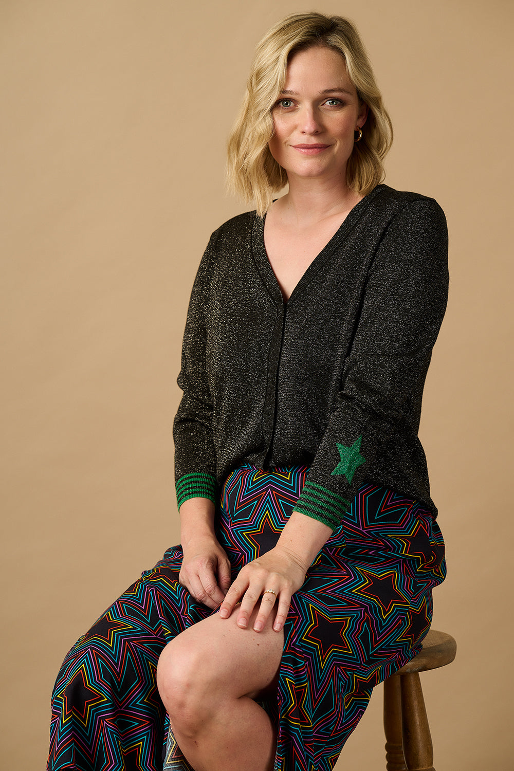 A woman sitting on a chair wearing a black long-sleeve cardigan with green stripes and a star cuff, paired with a colorful patterned skirt.