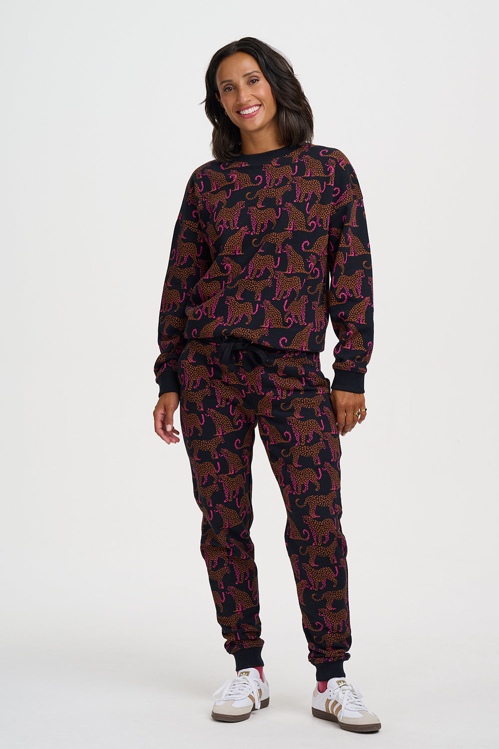 A woman modeling a matching two-piece loungewear set featuring a dark base with an allover animal print in brown and purple, paired with white sneakers against a plain light gray studio background.
