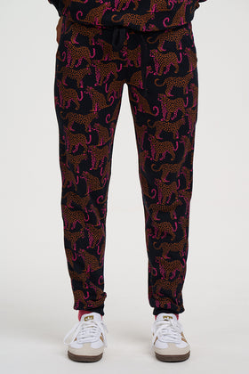 A person wearing black jogger-style pants with an allover leopard print in pink and orange, paired with white sneakers, shown from the waist down against a light gray studio background.