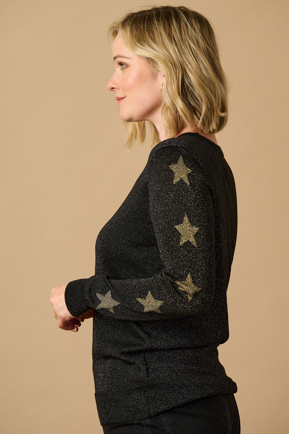 Woman wearing a black sweater with star patterns and black pants against a beige wall.