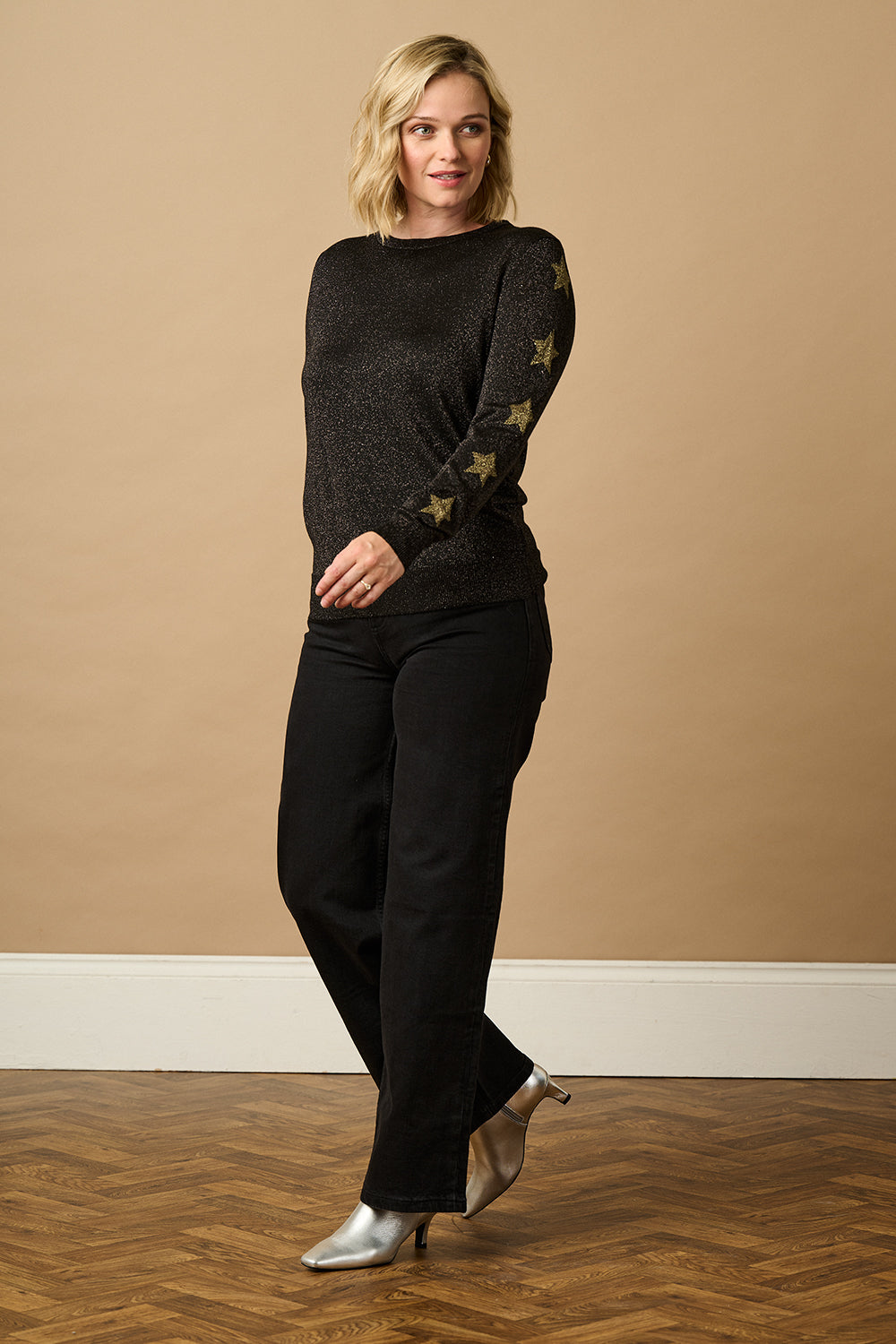 Woman wearing a black sweater with star patterns and black pants against a beige wall.