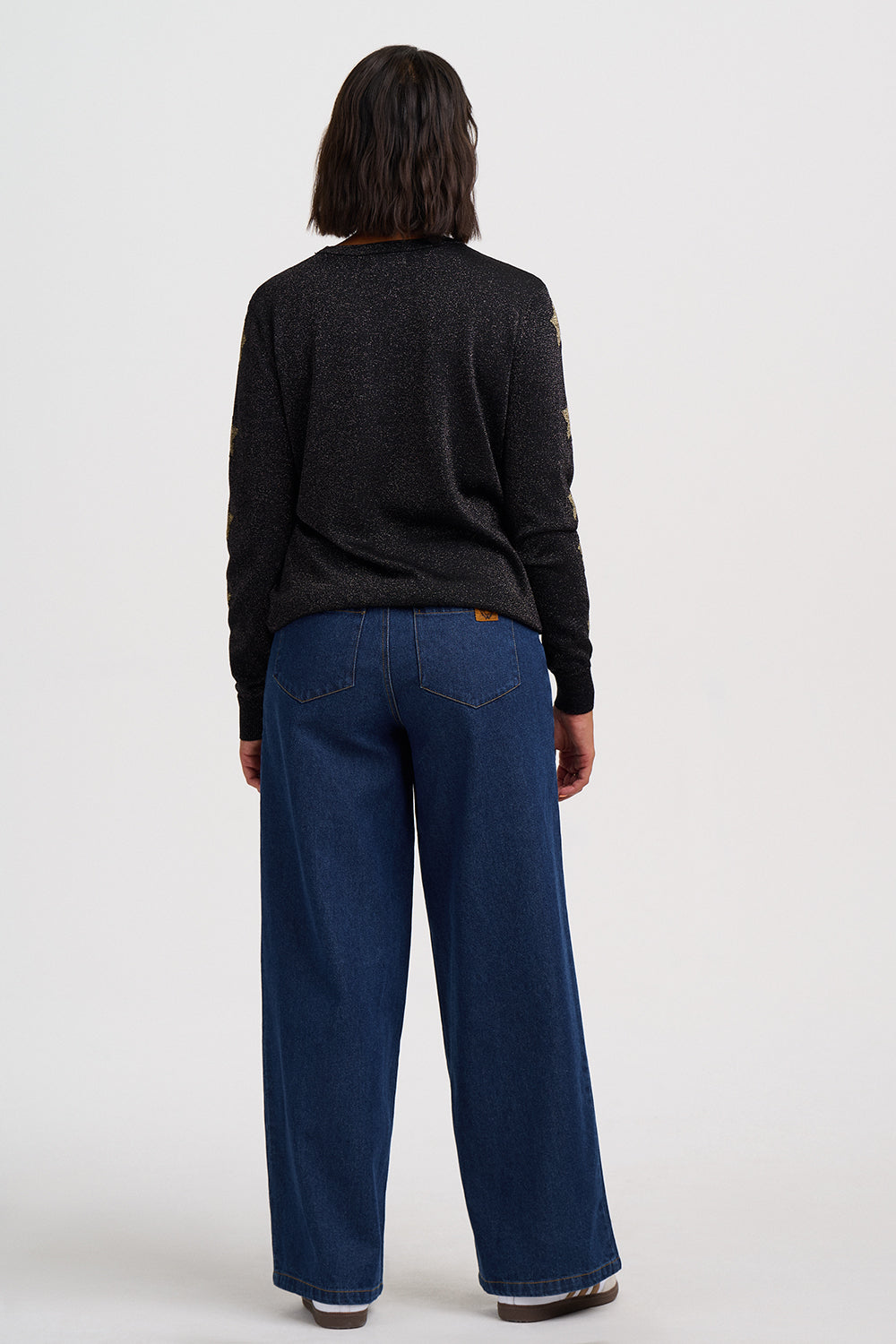 A model shown from the back wearing a dark charcoal knit sweater and dark blue wide-leg jeans, standing against a plain light gray studio background.