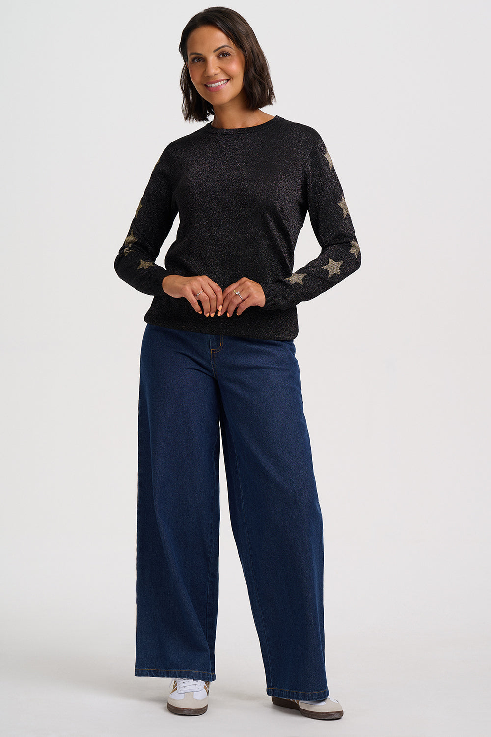 A woman modeling a black long-sleeve knit sweater with gold star patterns on the sleeves, paired with dark blue wide-leg jeans and white sneakers, against a light gray studio background.