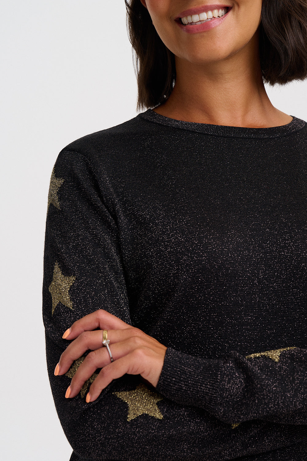 A close-up of a woman wearing a black knit sweater with metallic gold star patterns on the sleeves and hem, set against a plain light gray background.