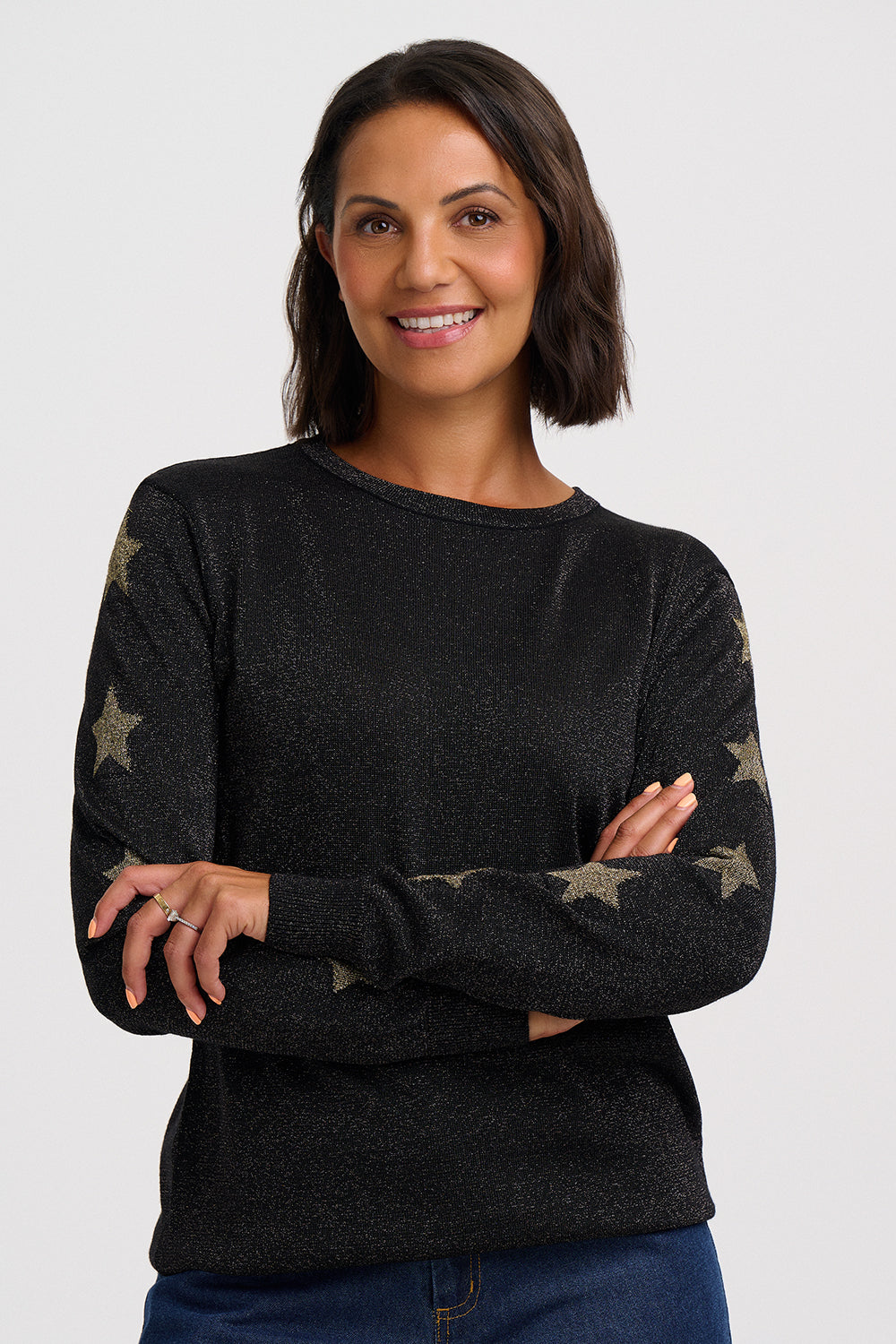 A woman with short dark brown hair smiling, wearing a black long-sleeve crewneck sweater with metallic gold star patterns on the sleeves, paired with blue jeans, against a light gray studio background.