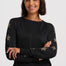 A woman with short dark brown hair smiling, wearing a black long-sleeve crewneck sweater with metallic gold star patterns on the sleeves, paired with blue jeans, against a light gray studio background.