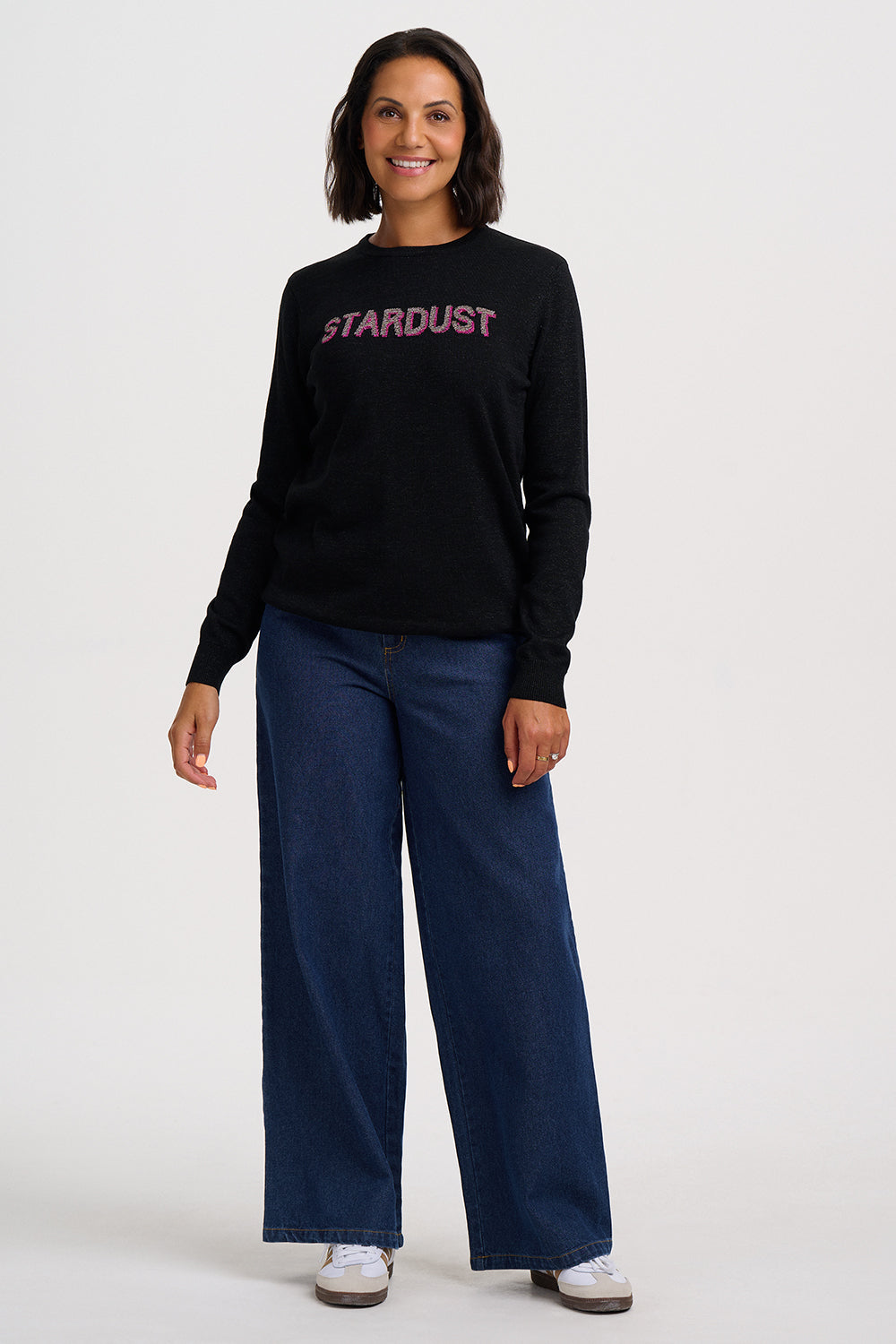 A woman modeling a black long-sleeve crewneck sweatshirt with 'STARDUST' printed in pink across the chest, paired with dark blue wide-leg jeans and white sneakers, against a plain light gray studio background.