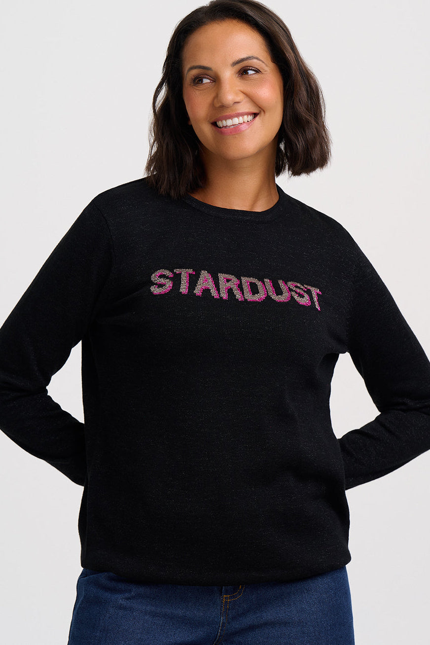 A woman wearing a black crewneck sweatshirt with 'STARDUST' printed across the chest in a textured pink font, paired with blue jeans, against a light gray studio background.