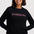 A woman wearing a black crewneck sweatshirt with 'STARDUST' printed across the chest in a textured pink font, paired with blue jeans, against a light gray studio background.