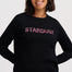 A woman wearing a black crewneck sweatshirt with 'STARDUST' printed across the chest in a textured pink font, paired with blue jeans, against a light gray studio background.